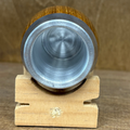 A close-up of a wooden and metal cylindrical object, possibly a small cup, tumbler, or tool component, resting on a small wooden display block against a textured brown background.